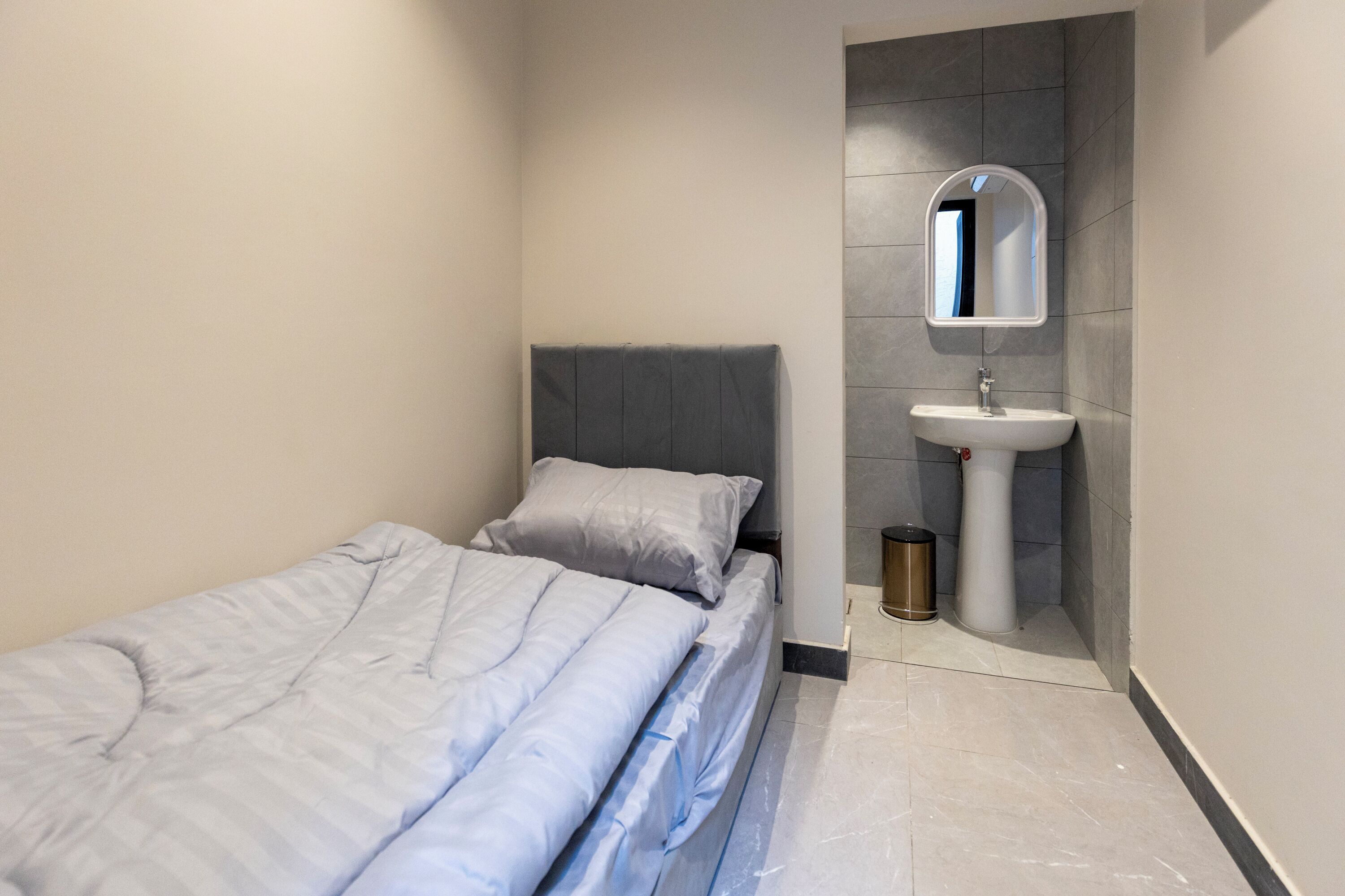 Une petite chambre à coucher moderne, avec un lit simple, une table de chevet, un miroir fixé au mur et une salle de bain comprenant un lavabo et des toilettes.