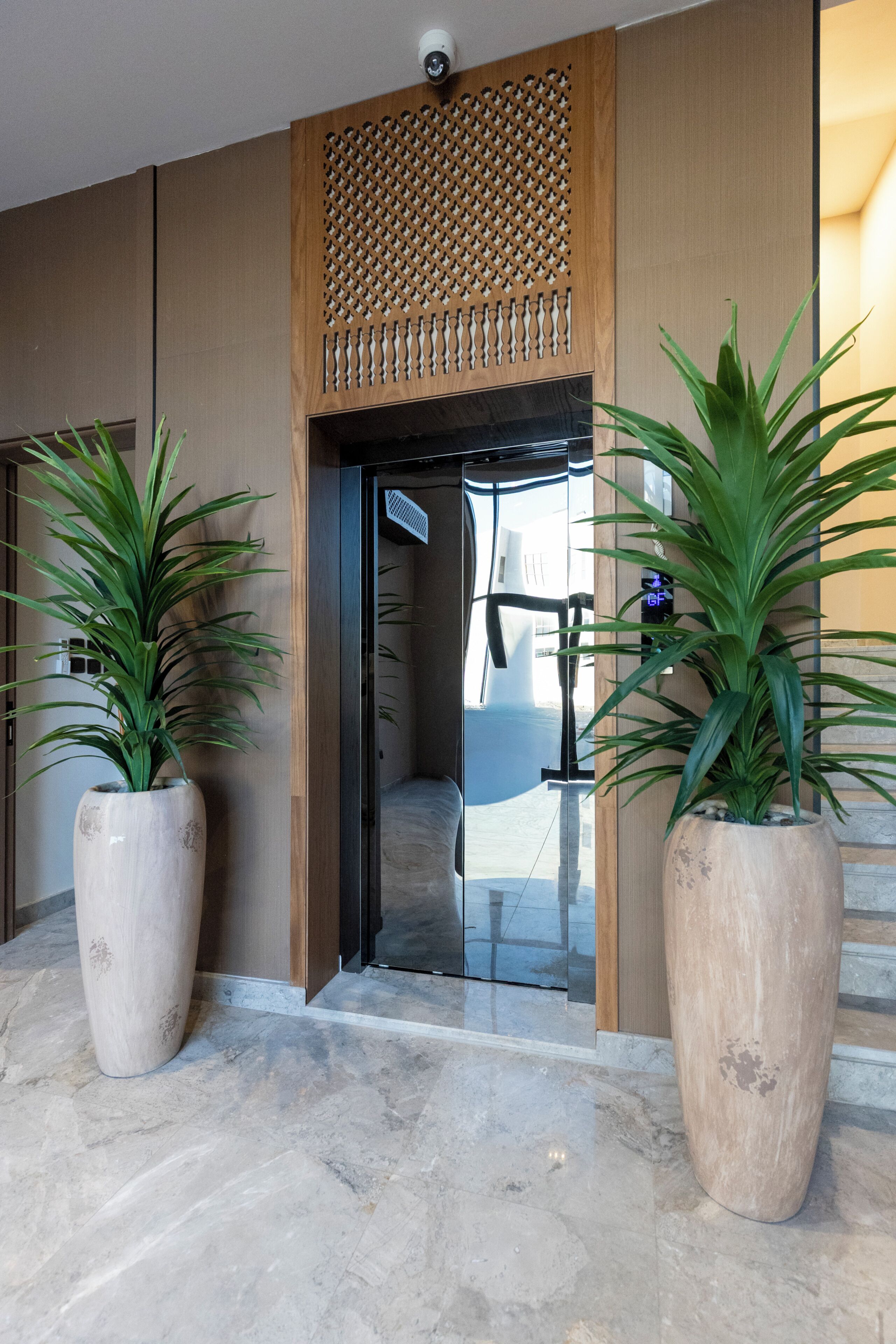 Un hall d’hôtel moderne, agrémenté d’un panneau décoratif en bois, de grandes plantes en pot et d’une porte en verre donnant sur l’intérieur.