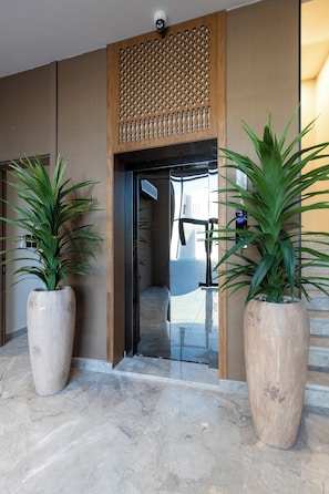 Um lobby moderno de hotel com um painel decorativo de madeira, plantas grandes em vasos e uma porta de vidro que dá para o interior.