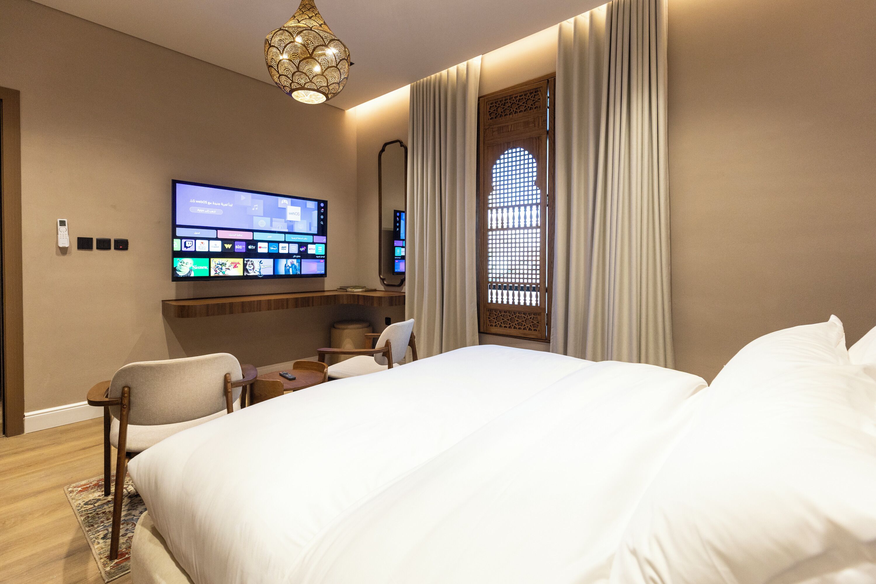 Ein Hotelzimmer mit einem großen Bett, einem flachbildfernseher an der Wand, einem Schreibtisch mit Stuhl und einem dekorativen Fenster mit Vorhängen.