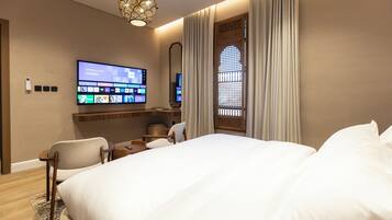 Ein Hotelzimmer mit einem großen Bett, einem flachbildfernseher an der Wand, einem Schreibtisch mit Stuhl und einem dekorativen Fenster mit Vorhängen.