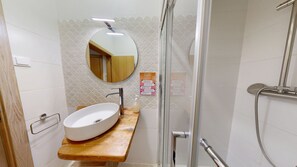 Un bany amb un mirall rodó, un lavabo de fusta, un rentamànega, un pavelló de dutxa de vidre i una barana.