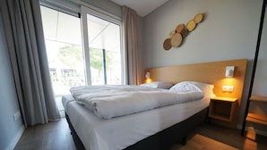 Ein Hotelzimmer mit Bett, Nachttisch, Wandleuchte und einem fensterbepflanzten Fenster.