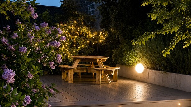 Una terraza de madera con una mesa de picnic y bancos, rodeada de follaje y luces colgantes.