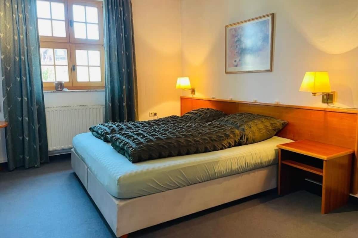 A bedroom with a bed, two bedside lamps, a window with curtains, a radiator, and a painting on the wall.