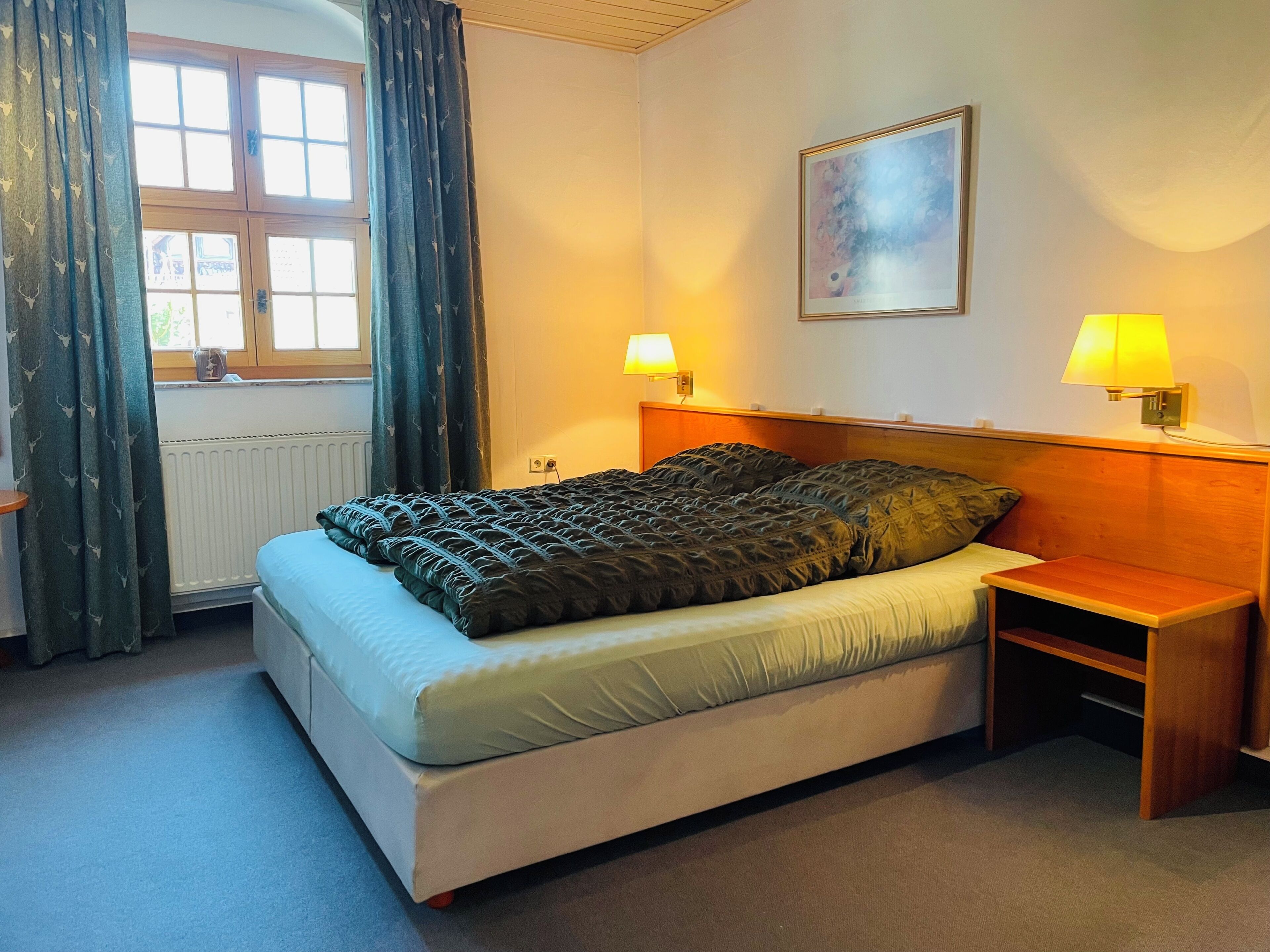A bedroom with a bed, two bedside lamps, a radiator, a window with curtains, and a painting on the wall.