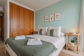 Cama bem arrumada com um cobertor verde, mesa de cabeceira com abajur e mesa de apoio em vime com uma planta.