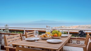 Taula de fusta amb plats, una testosseta i un bol de fruita a un balcó amb vista a una ciutat i muntanyes.