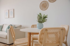 Uma área de refeições com cadeiras de vime, uma mesa redonda, um vaso com folhagem e um sofá com almofadas.