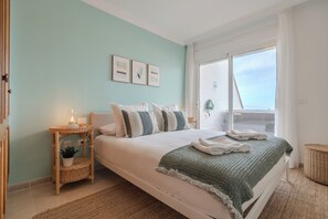 Ein Schlafzimmer mit Bett, Nachttisch mit Lampe, Fenster mit Vorhängen und Blick ins Freie.