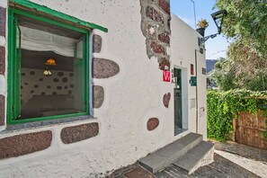A building with a window with a green frame and a white curtain, a door with a red sign, and a brick wall with holes.