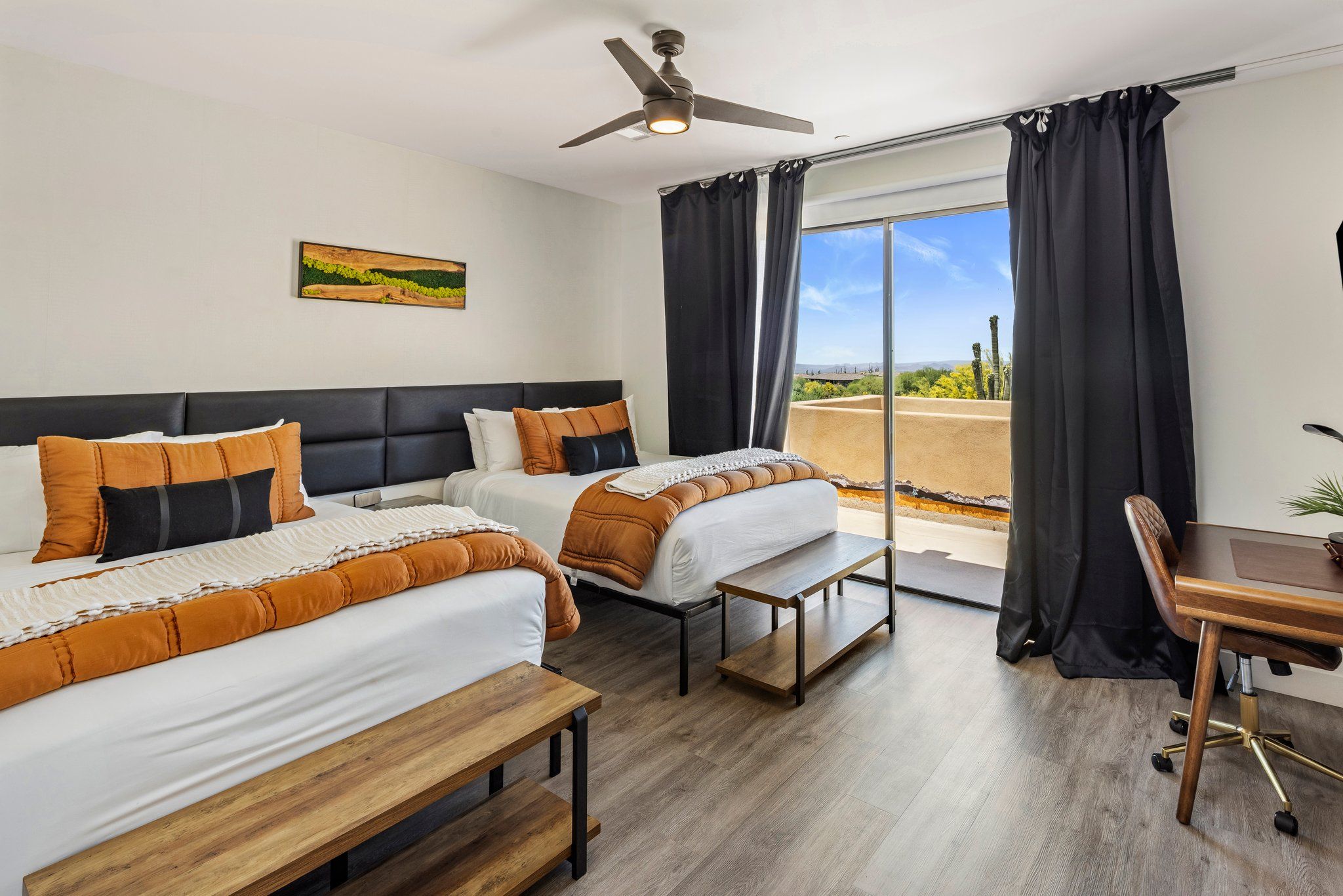 Ein Hotelzimmer mit zwei Betten, einem Schreibtisch, einem Stuhl, einem Deckenventilator und einem großen Fenster mit Vorhängen.