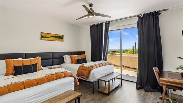 Ett hotellrum med två sängar, ett skrivbord, en stol, en takfläkt och ett stort fönster med gardiner.