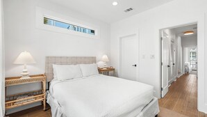 A bedroom with a large bed, two bedside tables with lamps, a wicker side table, a window with a view, and a door leading to another room. - NEW LISTING - Completely remodeled with new pics coming soon. (Seacrest Beach)