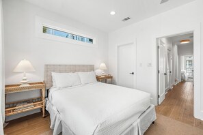 Un dormitorio con una cama grande, dos mesitas de noche con lámparas, una mesita auxiliar de mimbre, una ventana con vistas y una puerta que da a otra habitación.