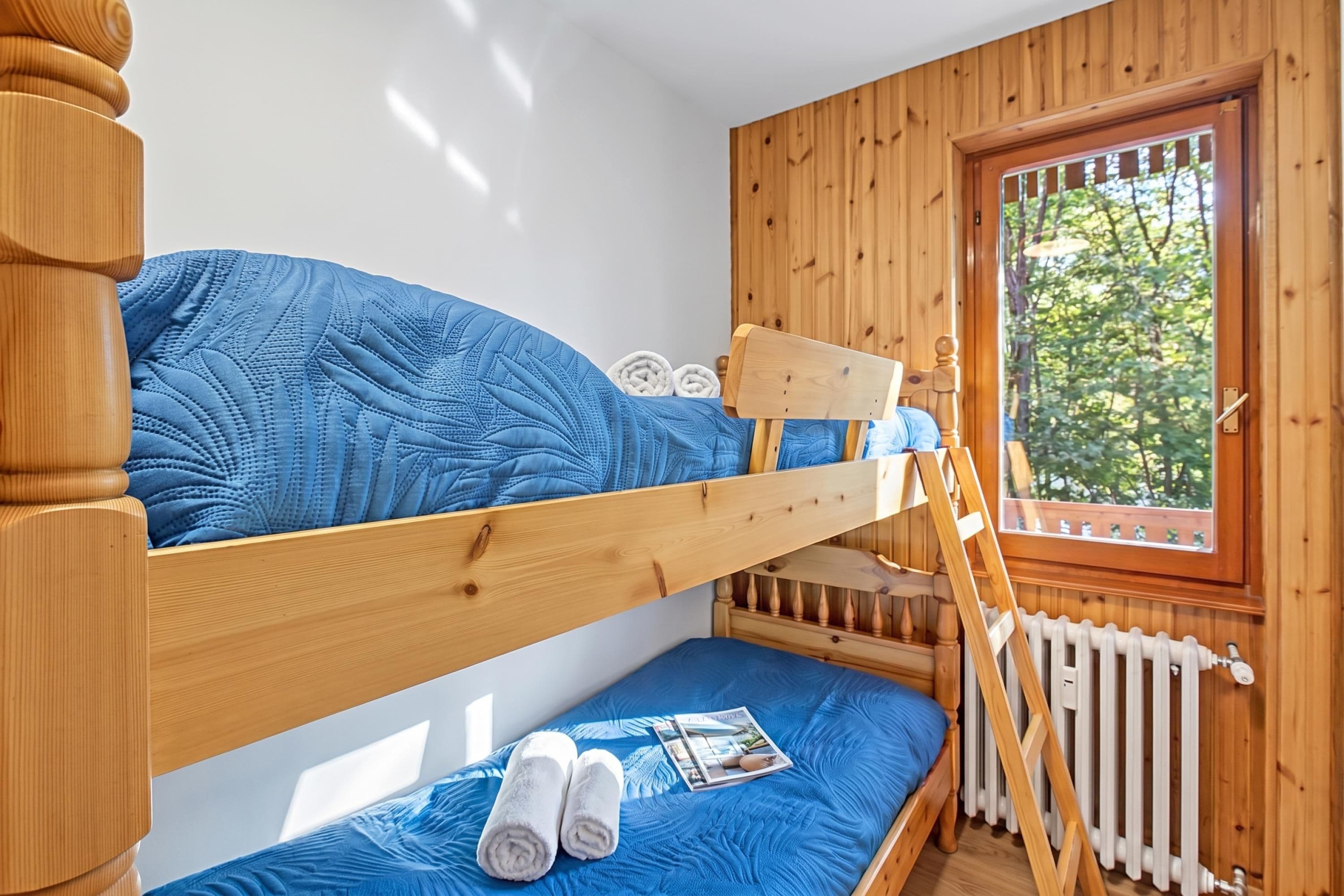 Habitación con literas, una escalera de madera, una ventana con vista a árboles y un radiador blanco.