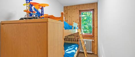 A bunk bed with a play structure on top, a wooden wardrobe, a white furry rug, and a window with a view of greenery.
