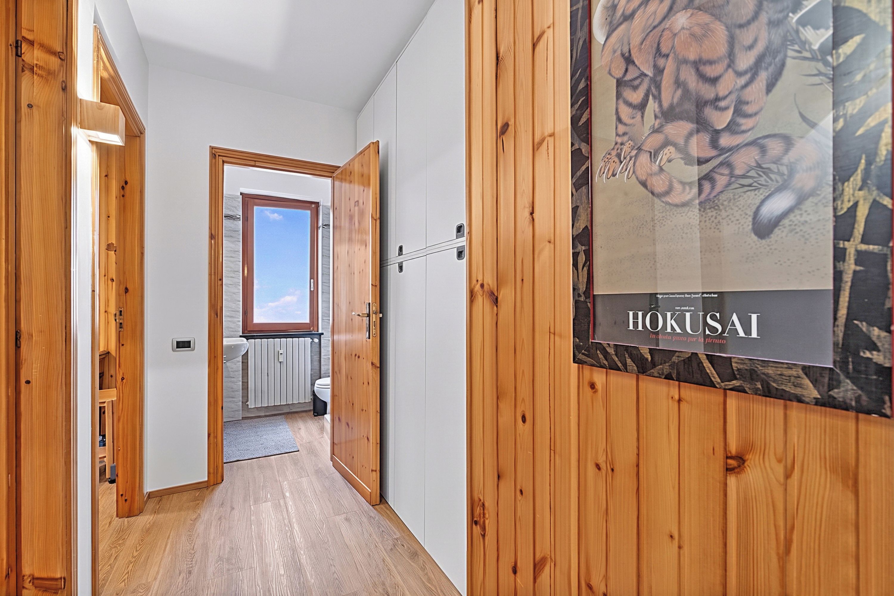 A hallway with wooden walls, a radiator, a window, and a door leading to a bathroom.