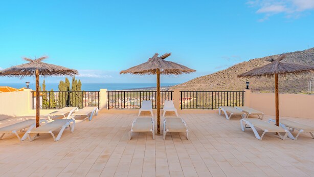 Eine Dachterrasse mit weißen Liegestühlen und Reetsonnenhüten, mit Blick auf eine Küstenlandschaft.