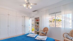 Ein Schlafzimmer mit einem großen Bett, einem Deckenventilator, weißen Schränken, einem Korbstuhl und Blick ins Freie.