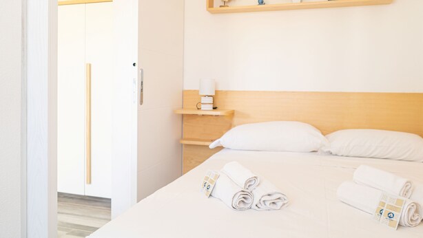 Una habitación de hotel con cama, almohadas, una mesita de noche, un estante con objetos decorativos y toallas enrolladas.