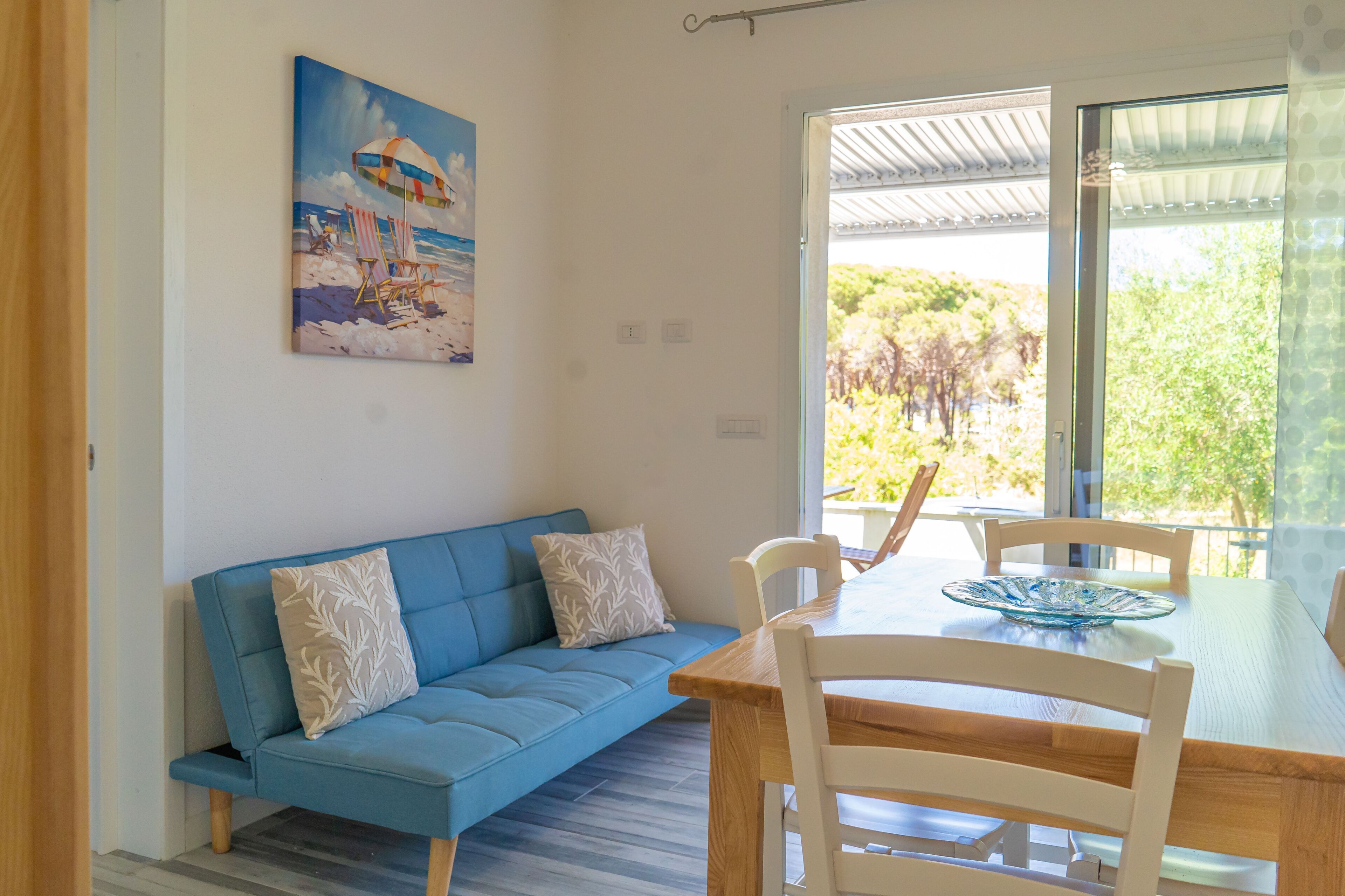 Une pièce avec un canapé bleu, une table à manger avec des chaises, un saladier en verre et un tableau représentant une scène de plage.