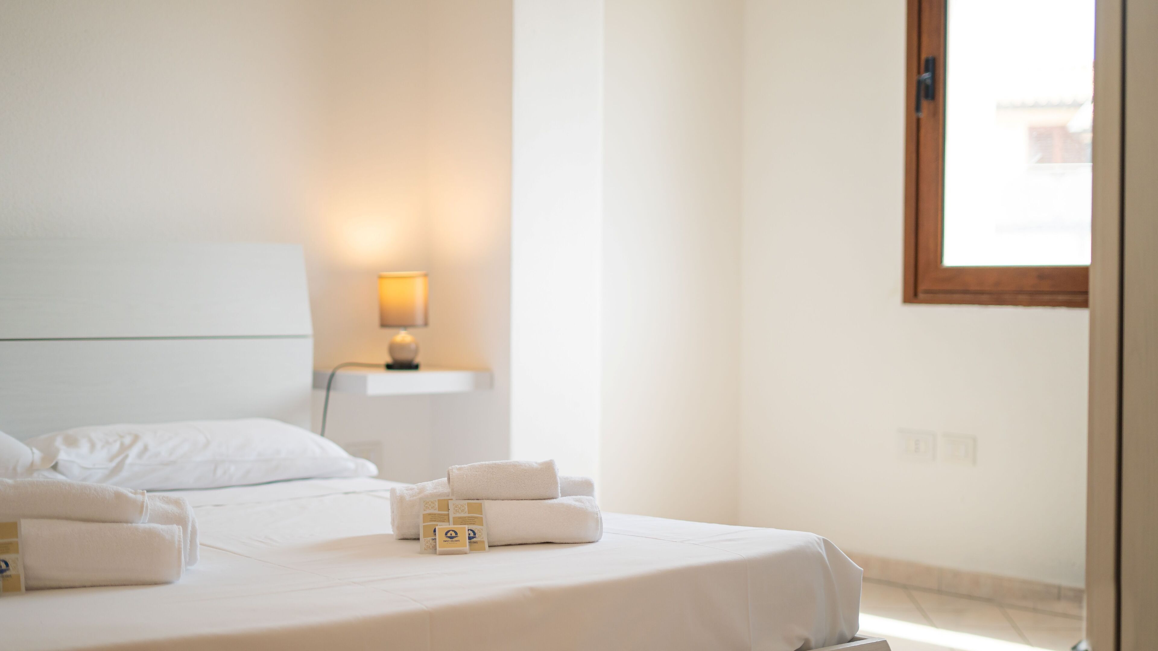 A hotel room with a bed, a nightstand with a lamp, and a window.