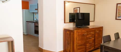Une pièce comprenant un meuble en bois, un téléviseur, une table à manger avec des chaises, et une cuisine à l’arrière-plan.