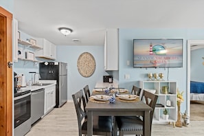 A modern kitchen with a dining area, a flat-screen TV, and a decorative wall piece. - Club Ocean Villa II 37 by Seaside Vacations & Sales (Ocean City)