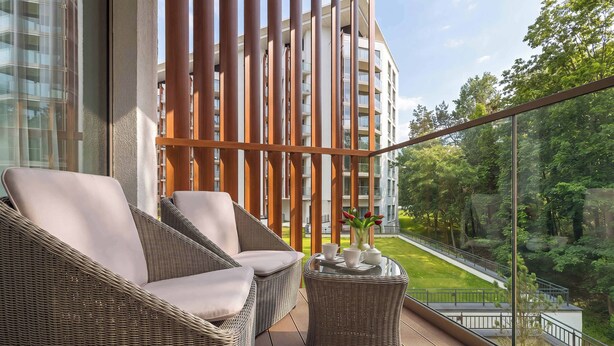 Un balcón con mobiliario de mimbre, una mesa de vidrio con tazas y un florero, y vistas a un edificio y follaje.