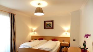 A hotel room with a wooden bed, two bedside lamps, a ceiling light, a framed picture on the wall, and a table with a potted plant. - Apartment 'Residence Katiuscia 5' with Mountain View, Private Garden and Wi-Fi (Selva di Val Gardena)