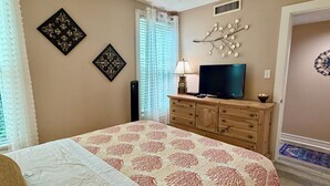 Quarto com cama, penteadeira, televisão, arte decorativa na parede e janela com cortinas.