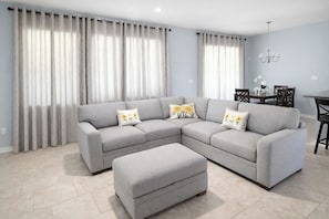 A living room with a grey sectional sofa, an ottoman, a dining table with chairs, and a chandelier. - Disney Oasis / Game Room, Pool & Screened Lanai (Haines City)