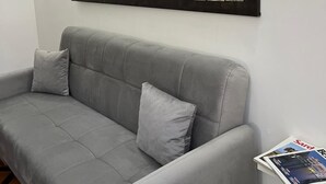 Un sofá gris con dos cojines, un cuadro con un mapa mundial en la pared, y una lámpara de pared con diseño de frutas.