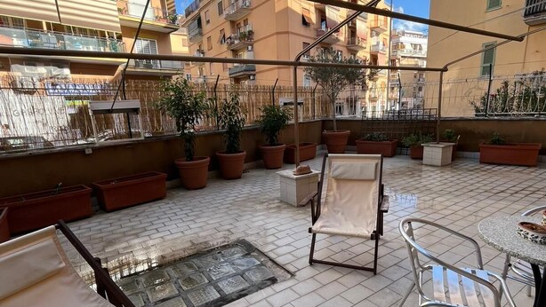Una terraza al aire libre con suelo de baldosas, sillas de metal, una mesa y plantas en macetas.