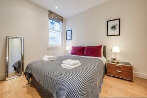 A neatly made bed with folded towels, a bedside table with a lamp, a framed picture on the wall, and a window with curtains. - Fitzrovia Apartments 3BR Sleeps 12 Oxford (London)