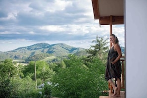 A person standing on a balcony with a view of green hills and a cloudy sky.