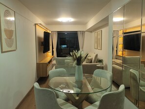 Een moderne woonkamer met een eettafel met glazen blad, comfortabele zithoek, een flatscreen televisie en een grote spiegel.