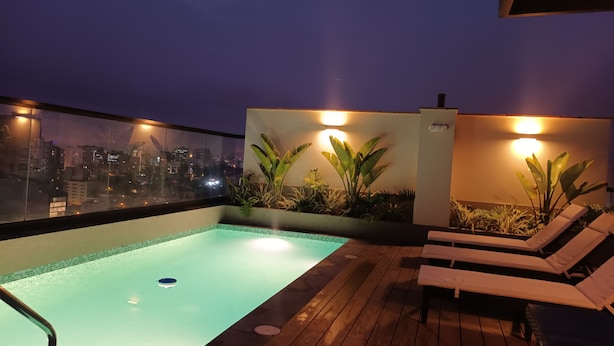 A rooftop pool with lounge chairs, plants, and city views at dusk.