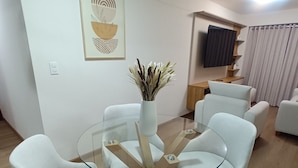 A modern living room with a glass-top coffee table surrounded by white armchairs, a wooden side table, and a wall-mounted TV.