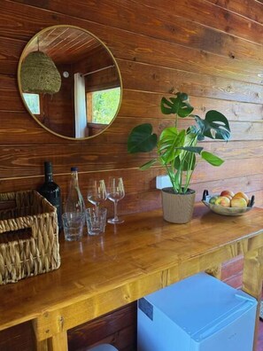 Una zona de bar con barra de madera, un espejo, una planta en maceta, una cesta y un bol de fruta.