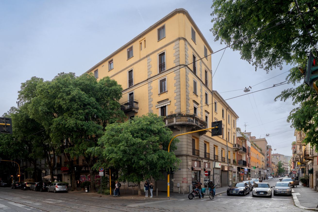 A multi-story building with balconies, parked cars, and trees along the street.
