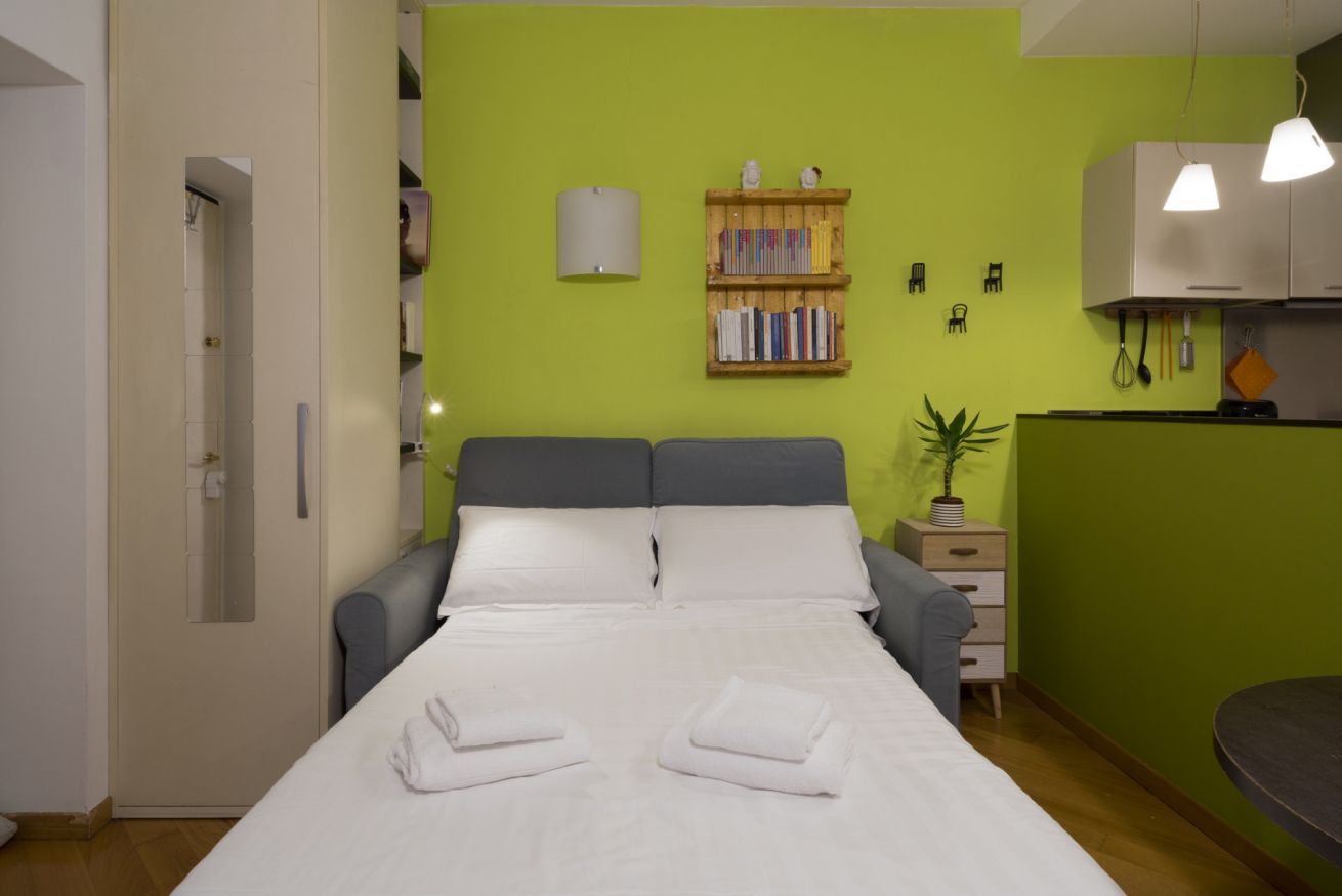 Een hotelkamer met een bed, nachtkastjes, een kleine plant, een boekenkast en een kitchenette.