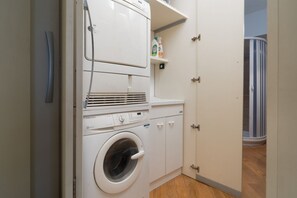 Ein kleiner Waschraum mit einer Waschmaschine und einem Trockner mit Frontlader, einem Regal für Reinigungsmittel und einer Tür zu einem anderen Raum.