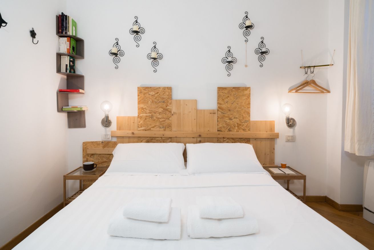 Una cama con sábanas blancas, dos mesitas de noche, un cabecero de madera, estanterías fijas con libros y apliques decorativos.