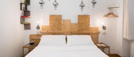 Una cama con sábanas blancas, dos mesitas de noche, un cabecero de madera, estanterías fijas con libros y apliques decorativos.