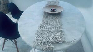 Ein runder Tisch mit einer Wanddekoration aus Makramee, zwei schwarze Stühle und Blick auf das Meer und Gebäude.
