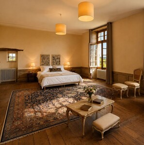 Un dormitorio con una cama grande, dos lámparas de noche, una ventana con cortinas, suelo de madera y una pequeña mesa con un libro.