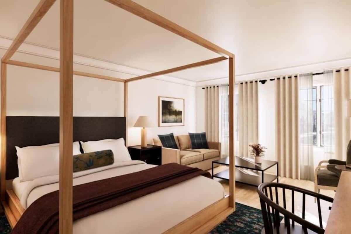 Hotellihuoneessa on suuri sänky, jossa on neljä pylvästä ja tumma pääty, sohva, pieni pöytä ja tuoli. Huoneessa on suuret ikkunat ja verhot.
