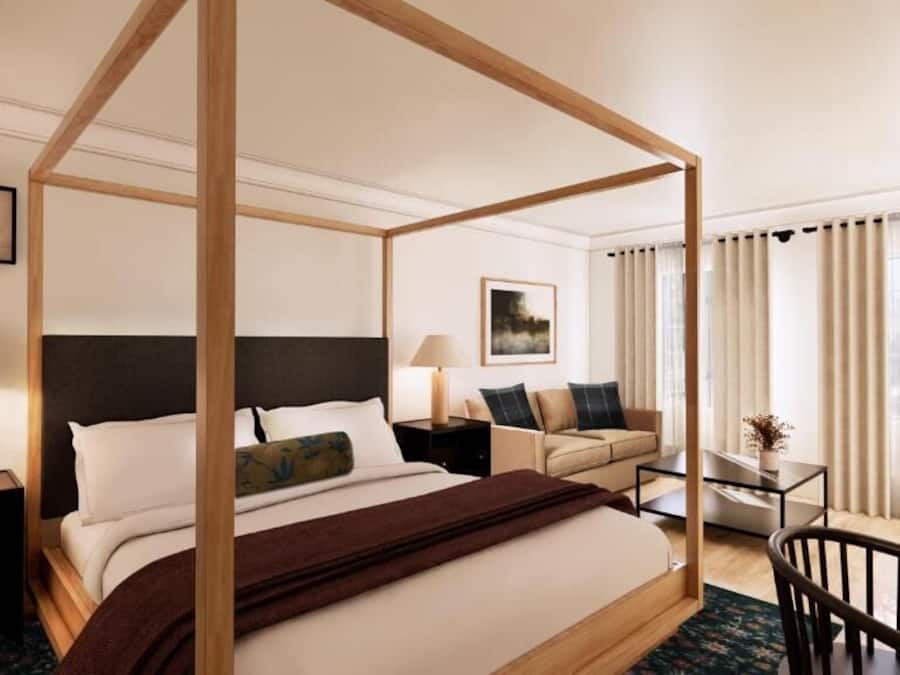 A four-poster bed with a dark headboard, a sofa, a small table, and a chair in a hotel room with large windows and curtains.